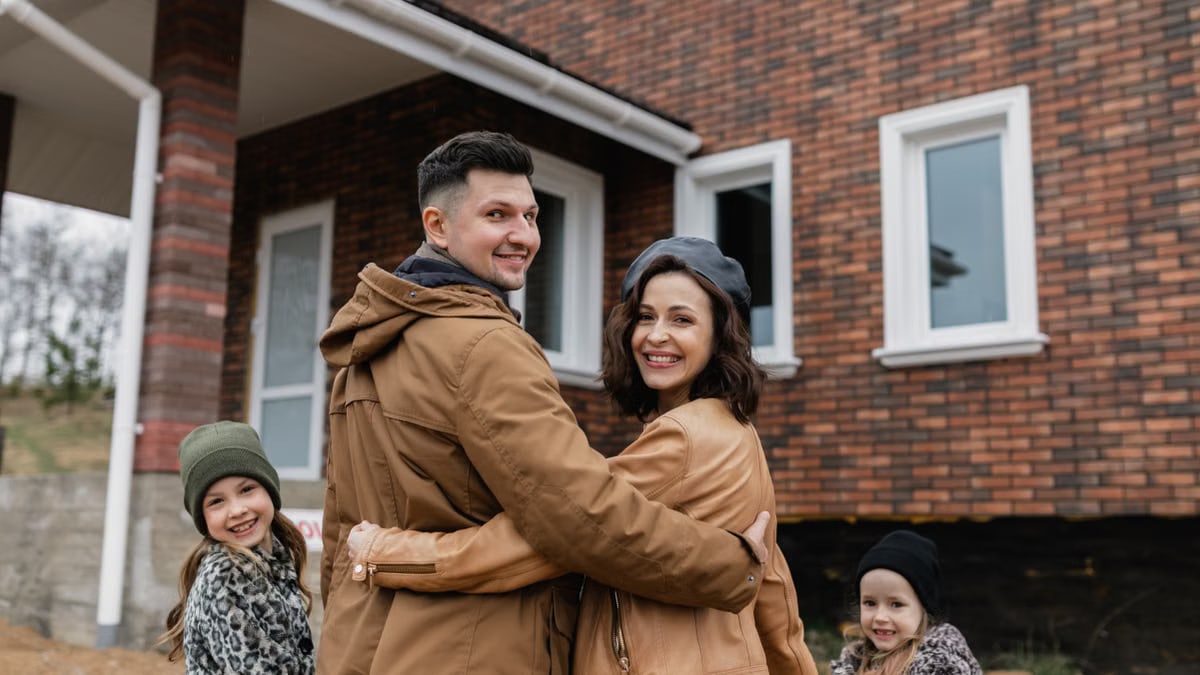 Family of four happily posing in front of their new brick house on a sunny day.
